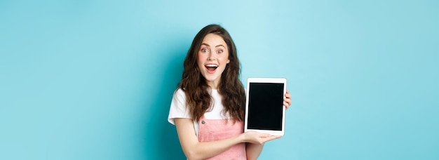 面白がってかわいい女の子は空白のデジタル タブレット画面を示し、畏敬の念を示すカメラで興奮して笑顔