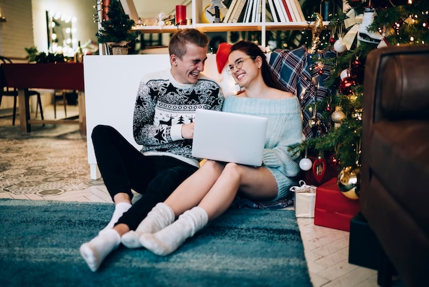 Amused young couple browsing on laptop beside Christmas tree