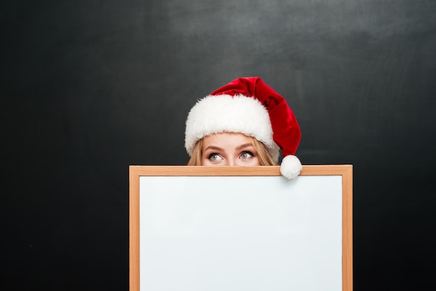 Amusing shy girl in santa claus hat hiding behind blank white board