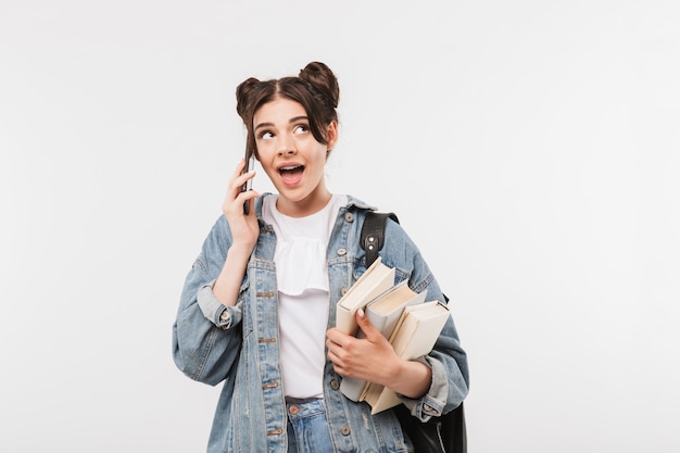 amusing student girl with double buns hairstyle wearing jeans clothing and backpack talking on smartphone while holding studying books, isolated on white wall