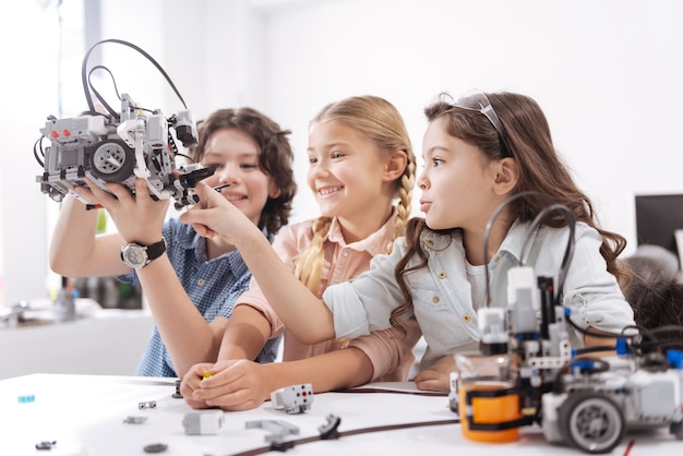 Amusing with our electronic friend. Positive happy joyful pupils sitting at school and playing with robot while working on the tech project