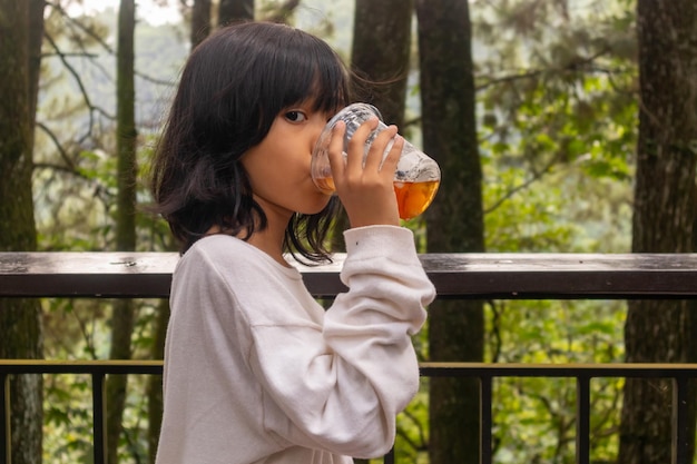 写真 自然の木の背景のバルコニーでプラスチックのカップのアイスティーを飲んでいるアジアの小さな女の子