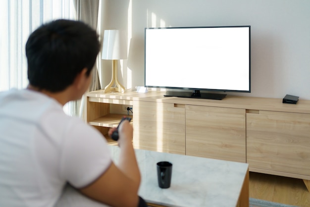 写真 アジア人の男性がテレビのリモコンを持っており、自宅の居間のソファでテレビを見ながらチャンネルを押しています。