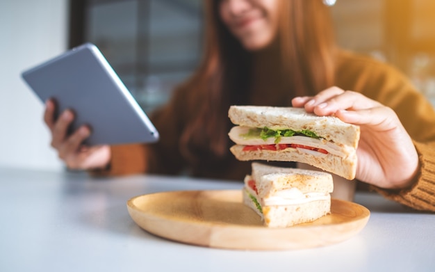 タブレットpcを使用しながら全粒小麦のサンドイッチを持って食べているアジアの女性