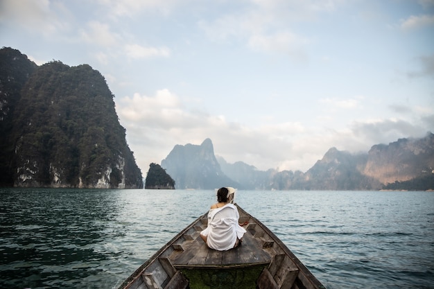 写真 白いシャツを着たアジア人女性が、タイ旅行中に海の真ん中に美しい山があるボートの前に座っています。素晴らしいタイ