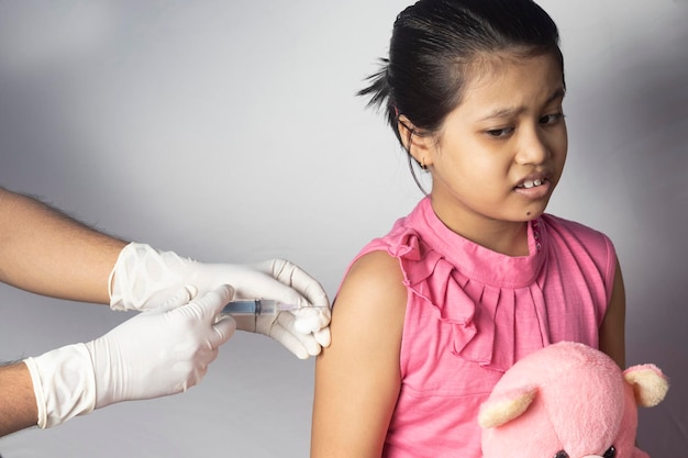 Фото Индийская девочка с плюшевым медведем на коленях получает дозу вакцины на белом фоне
