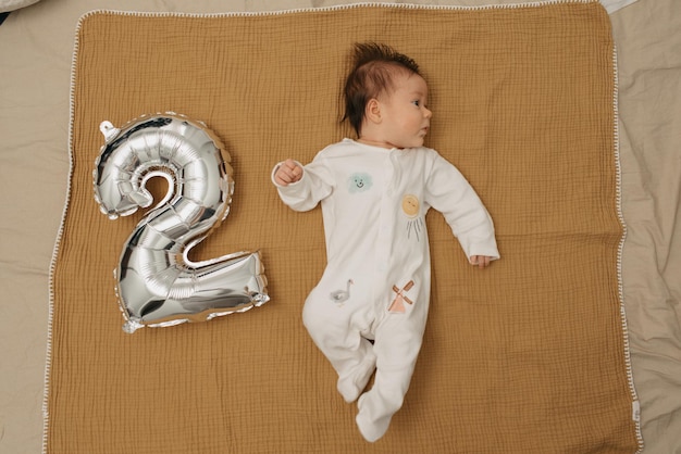 写真 乳児は、2の形をした銀箔の風船の近くのモスリン毛布でリラックスし、右を見つめています。ワンピースの女の赤ちゃんが2ヶ月を祝っています。