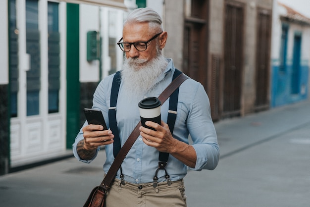 写真 流行に敏感な服と眼鏡と長い白ひげを着た年配の男性が通りを歩きながらコーヒーを飲み、彼の携帯電話を見ます