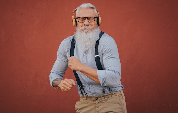 Фото Пожилой мужчина в хипстерской одежде и очках с длинной белой бородой слушает музыку и радостно танцует на улице.