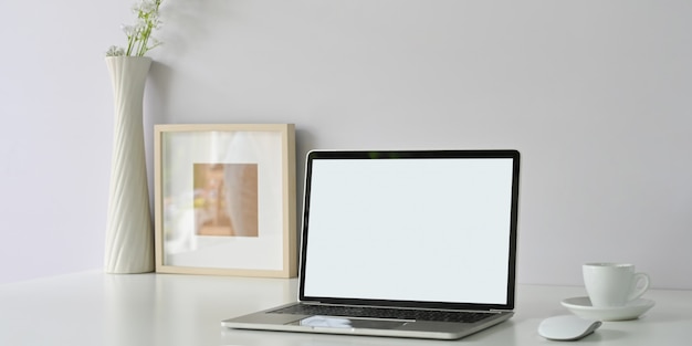 整然としたワークスペースは、白い空白の画面のコンピューターラップトップ、額縁、コーヒーカップ、花瓶に囲まれています。