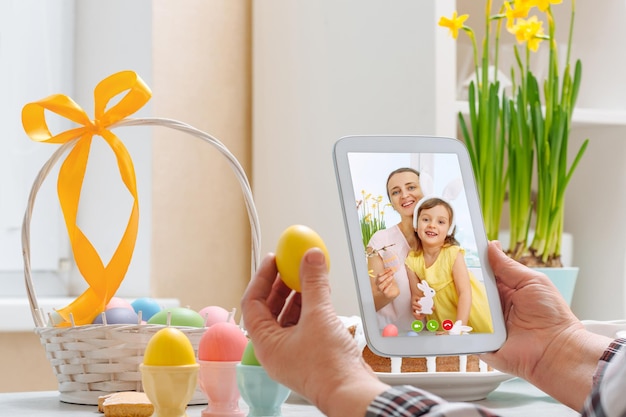 Фото Женщина держит в руках цифровой планшет и желтое яйцо и разговаривает по видеосвязи в пасхальные праздники.