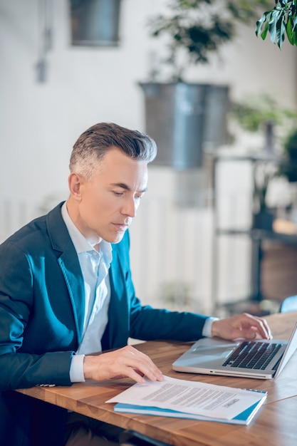 Analyse. Attente betrokken man in licht shirt en donkere jas die document analyseert achter een laptop die op kantoor werkt