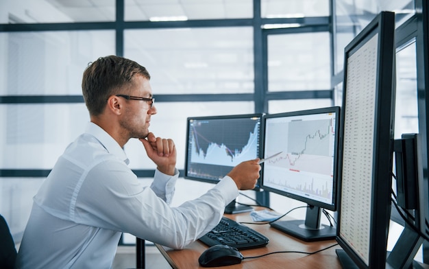 Analysing graphs. Young businessman in formal clothes is in office with multiple screens. Conception of exchange and money.