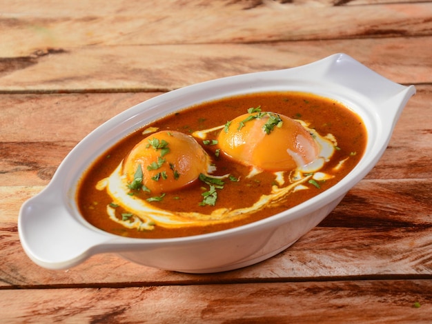 안다 마살라(Anda Masala) 또는 에그 카레(Egg Curry)는 소박한 나무 배경 위에 세라믹 그릇에 담긴 인기 있는 인도 매운 음식입니다.