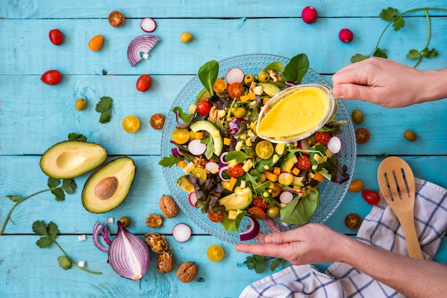 ands of a woman pouring a yellow sauce over a fresh salad on a blue background