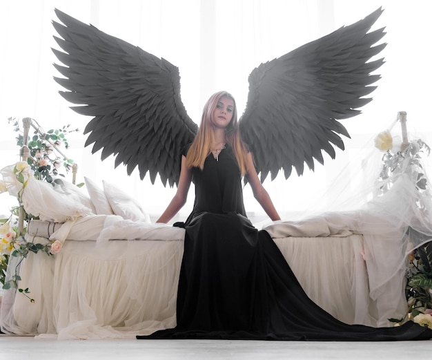 An angel with black wings on a white bed in flowers A young thoughtful girl is sitting on the edge of the bed