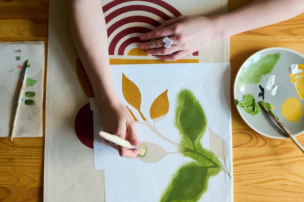 Above angle of hands of young artist with paintbrush painting green leaves