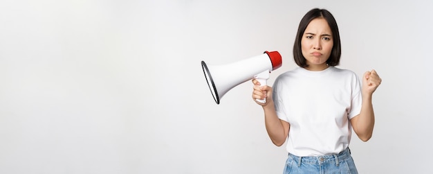 Angry asian girl activist holding megaphone and looking furious protesting standing over white backg...