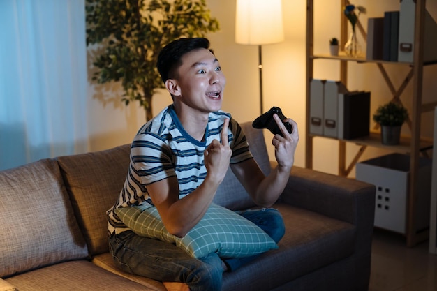 Angry asian japanese man in casual with joystick in hands and
point middle finger to tv monitor. mad guy gamer lose and yelling
at television screen in night home living room. male on sofa
depressed.