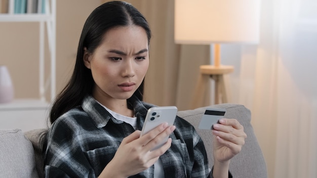 Angry asian woman sit on sofa at home hold credit card use mobile phone try to pay with ebank app