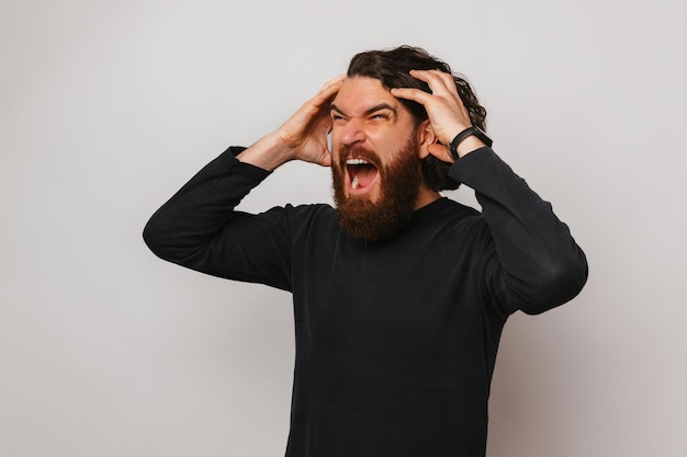Angry bearded man is screaming and holding his head because of headache
