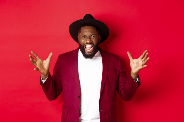 Angry Black man screaming and shaking hands with hatred, losing temper, standing outraged against red background