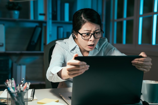 Angry businesswoman looking holding attentively at laptop computer screen mad about error of technology. stressed female freelancer worried about deadline doing remote job overtime at evening office