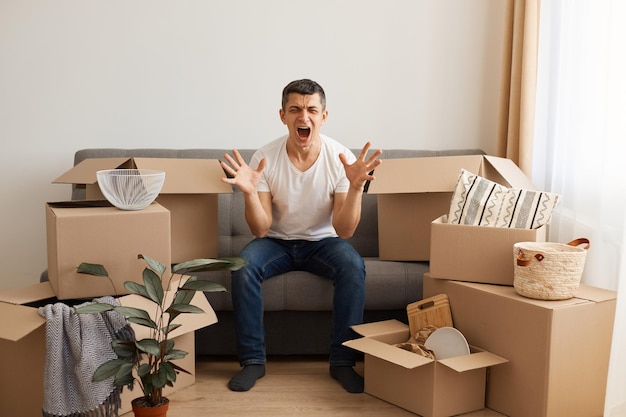 Angry crazy man wearing white T-shirt and jeans sitting on sofa surrounded with pile of packages, moving to a new apartment, doesn't want to unpack boxes with belongings, screaming with anger.