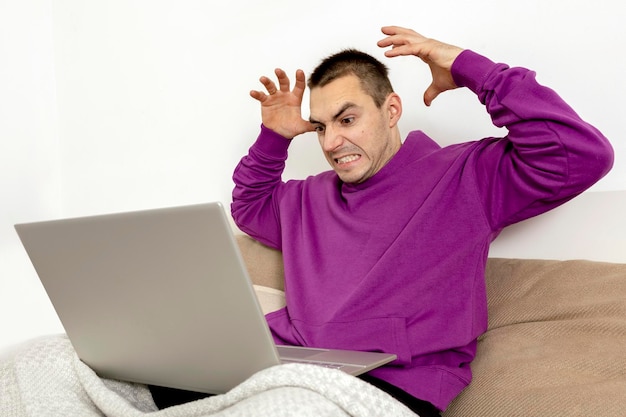 Angry and dissatisfied man with violet hoodie sitting on bed with laptop computer. Man using notebook to surf in internet, read news, watch movie, study or work online. Negative emotions, stress.