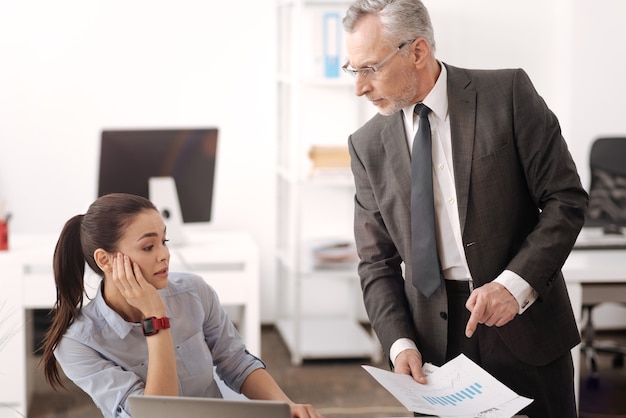 Angry elderly man keeping documents in right hand while giving new task, standing near his worker