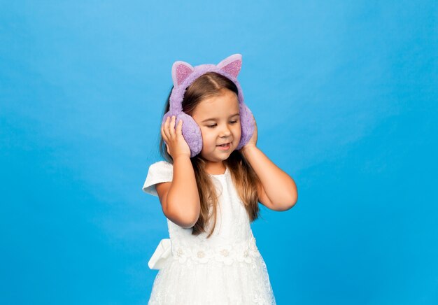 Angry emotional little girl in fur headphones on a blue background.