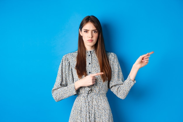 Photo angry frowning woman pointing fingers right and condemn person, feeling disapointed, standing in dress on blue.