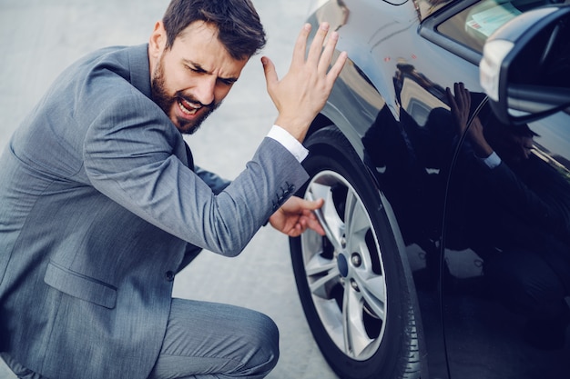 Angry handsome Caucasian businessman crouching next to his car, yelling and swearing. Flat tire.