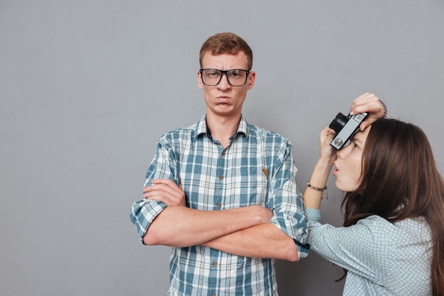 Angry hipster man in eyeglasses standing with arms folded while being portraitgraphed by a woman isolated on gray