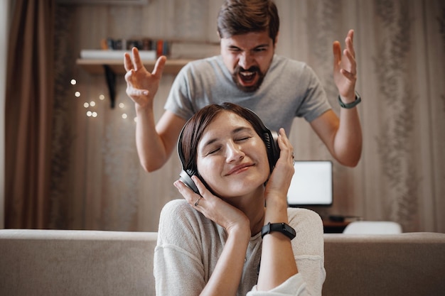 Angry husband shouting at wife sitting on sofa and covering ears with headphones