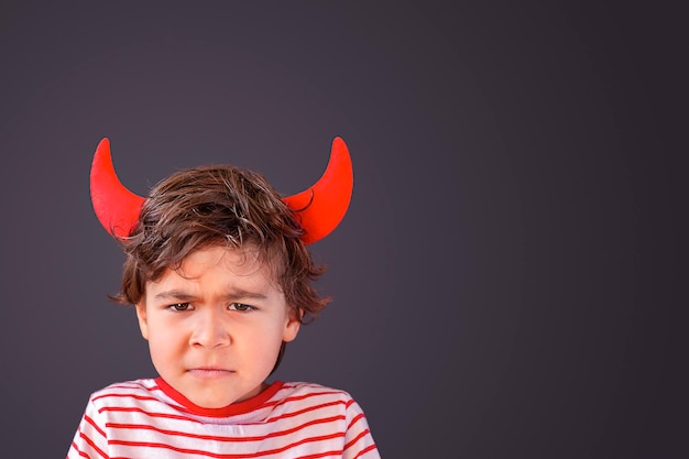 Angry little boy with devil horns on his head against grey background