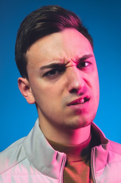 Angry, looking on. Close up caucasian man's portrait isolated on blue  wall in pink neon light. Beautiful male model. Concept of human emotions, facial expression Copyspace.