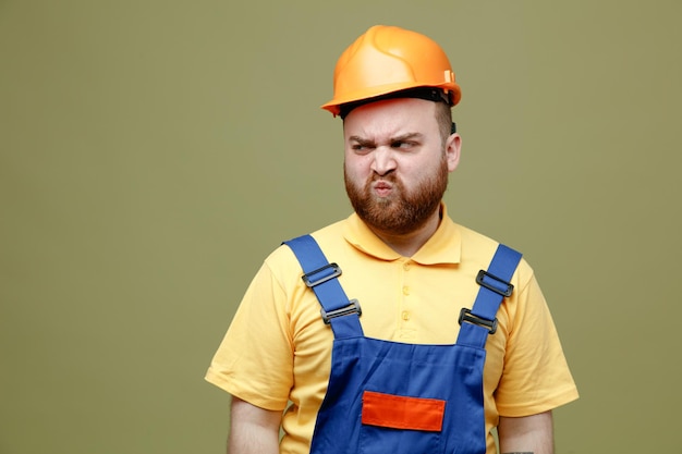 Angry looking at side young builder man in uniform isolated on green background