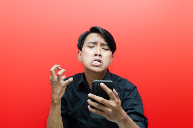 angry mad screaming asian man with mobile phone isolated over red background.