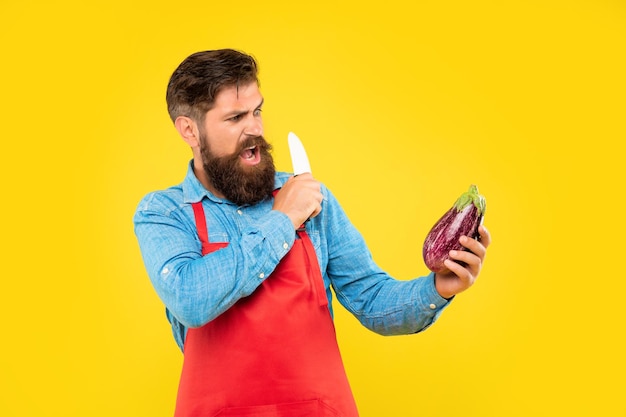 Angry man in apron holding cooks knife and eggplant yellow background cook