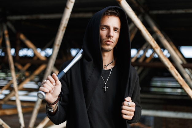 Angry man in black clothes holding a bat on the of metal pipes