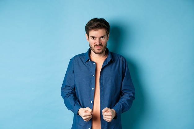 Angry and pissed-off man clench fists and stare with disdain at camera, trying to keep himself together, look with rage, standing on blue background.