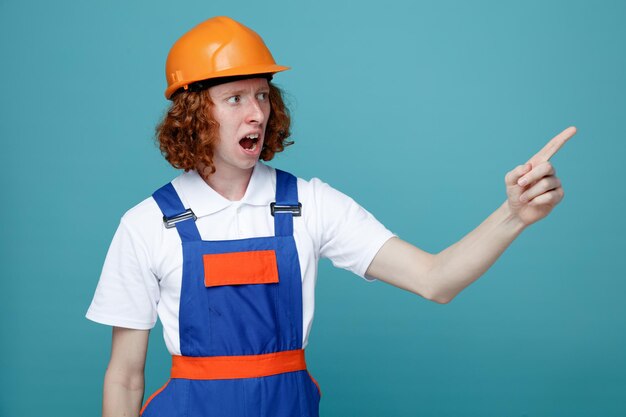 Angry points at side young builder man in uniform isolated on blue background