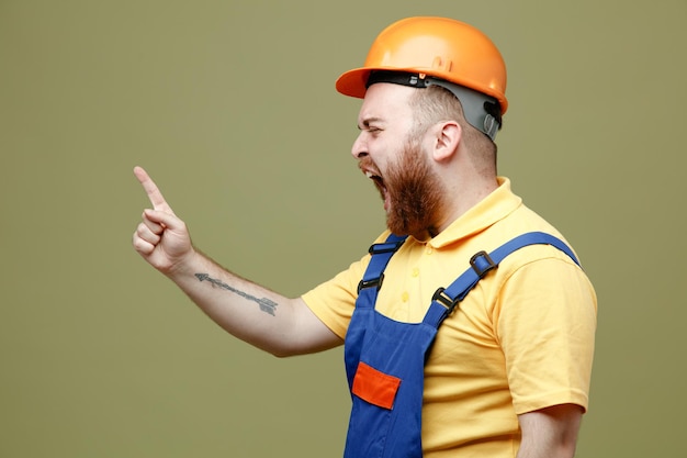 Angry points at side young builder man in uniform isolated on green background