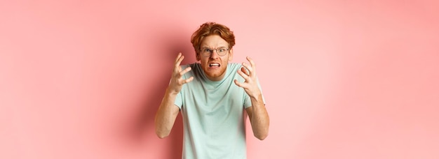 Foto ragazzo rosso arrabbiato con gli occhiali che urla, accigliato e stringe la mano a una persona frustrata e indignata.