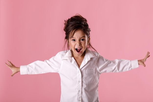 Angry screaming little child girl in white shirt with hairstyle\
looking to camera on pink background. human emotions and facial\
expression