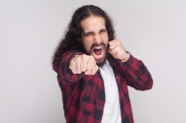 Angry screaming man with beard and black long curly hair in casual checkered red shirt standing, looking at camera and attacking with boxing fists . indoor studio shot, isolated on grey background