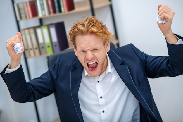 Angry screaming redhaired young man with closed eyes and raised fists in business suit at office