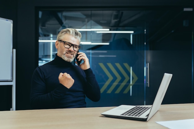 Angry senior businessman sitting in the office at the table with a laptop and worriedly talking on