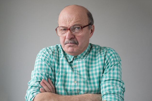 Angry senior man in glasses crossing arms looking annoyed at camera